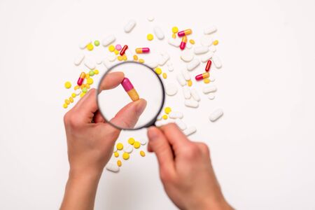 Hand is holding magnifier. Look through a magnifying glass at different pills on white background. Selective focus. Health, medicine and science concept. Top viewの写真素材