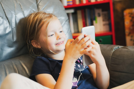 Portrait of cute blond hair little girl holding and playing smartphone indoor at home, close up with...