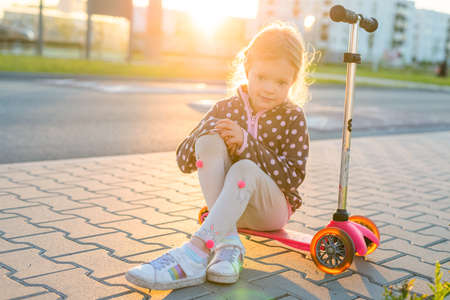 Outdoor portrait of cute blonde hair little girl sit on kick scooter on a road with sunny blurred backgroundの写真素材