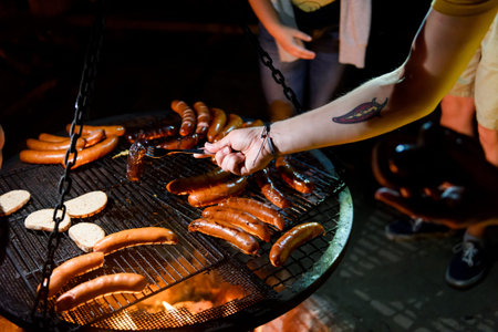 Making barbeque at night. Human hand taking grilled sausage from outdoor grill. Open air camp