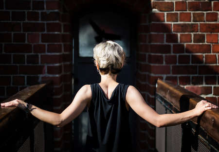blond hair woman in a black dress standing with her back on a bridge against with brick wall. Young stylish woman walking on the city streetの写真素材