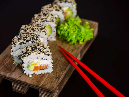 Uramaki Sushi Roll with salmon fish, avocado and cream cheese philadelphia, sushi roll covered with sesame seeds on wooden board on black backgroundの写真素材