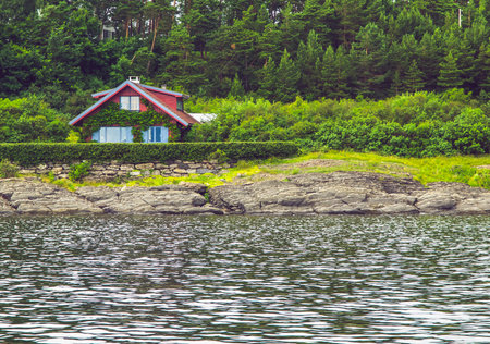 Red cozy wooden house covered with ivy on a oslofjord rocky shore. Norwegian fjords landscapeの写真素材