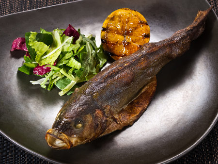 baked trout on a plate with grilled lemon and salad mix on dark background, horizontal photo