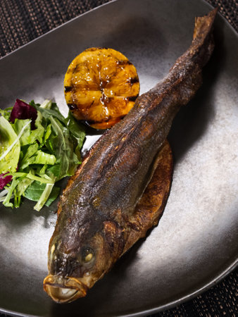 baked trout on a plate with grilled lemon and salad mix on dark background, vertical photoの写真素材