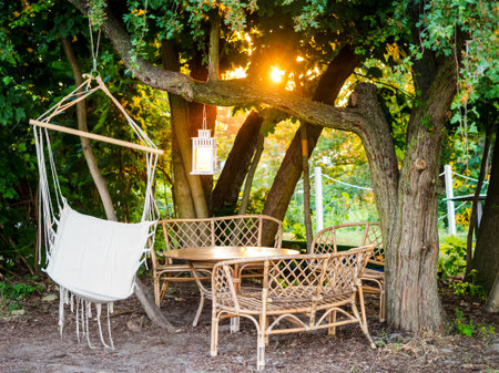 Wooden garden patio with wicker furniture chairs, table and a hammock, green trees in a summer setting on a sunset. garden seating area. Cozy hygge place for weekend relax in gardenの写真素材