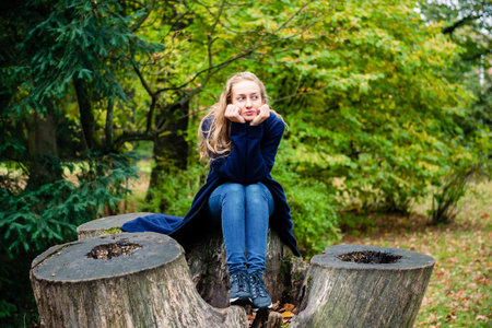 pretty blond female wearing long hoodie is sitting on a stump and looking side, outdoor in the city park in fall seasonの写真素材
