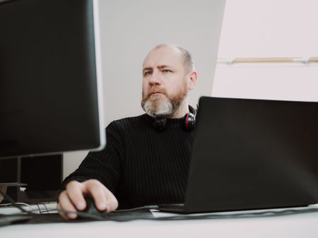 Adult focused bearded man remote working on laptop and using two monitors from home