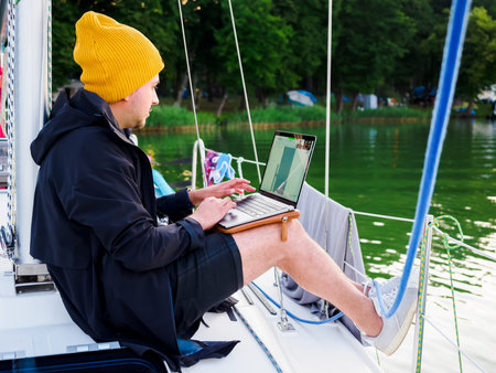 handsome young person wear jacket and knitted hat working with laptop on a sailing yacht during...