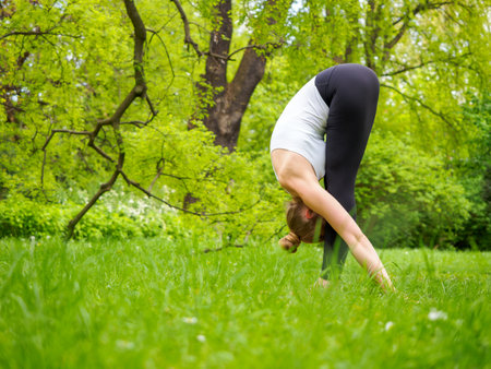 young pretty blond woman practicing yoga outdoor in the park in a sunny summer day, standing in padangusthasana poseの写真素材
