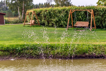 Two wooden swing benches in the garden with water drops in the foreground. Fountain water drops in the air with wooden garden swings in the background. Country summer view.の写真素材
