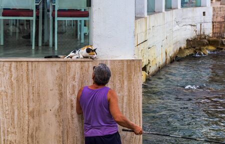 Valletta, Malta - August 30, 2019: Maltese fisherman patting stray calico cat while catching the fish with rod in another hand. Friendship of man and cat. Man caring for street cats.のeditorial素材