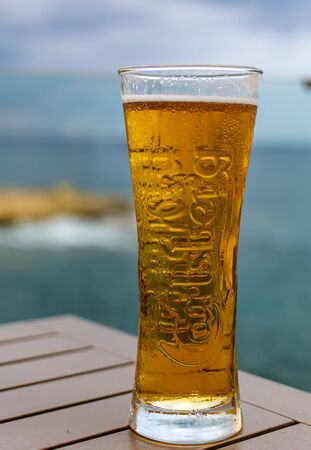 Sliema, Malta - September 5, 2019: Glass of light Carlsberg beer on wooden table with blurred sea on background. Evening relaxation.のeditorial素材