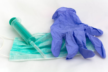Used green disposable face mask, blue latex glove and plastic syringe lying on a white table....