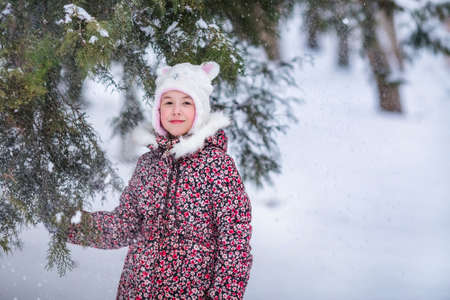 Smiling girl with white fur hat like a cat. Winter snowy background and gteen tree.の写真素材