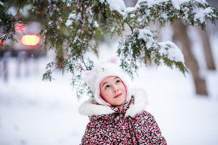 Portrait of smiling girl with white fur hat like a cat. Winter snowy background and gteen trees.