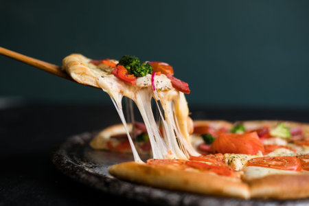 Delicious cheese stringy slice lifted of full supreme pizza baked fresh out of the oven next to ingredientsの写真素材