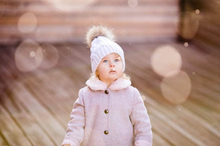 Portrait of cute little baby girl in pink coat and knitting hat with fur. Bokeh and unfocused snow...