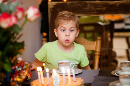 Beautiful adorable six year old boy in green shirt, celebrating his birthday, blowing candles on homemade baked cake, indoor.の写真素材