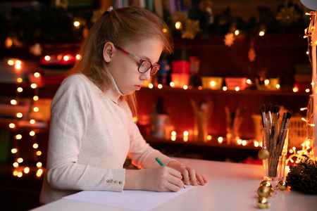 Blonde child girl with big pink ans black glasses writing letter to the Santa Claus or drawing something on the background with warm yellow bokeh. Christmas and New Year theme.の写真素材
