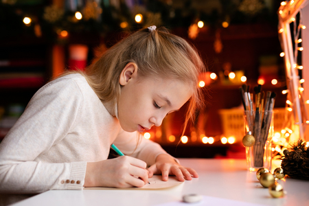 Blonde child girl writing letter to the Santa Claus or drawing something on the background with warm yellow bokeh. Christmas and New Year theme.の写真素材