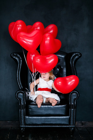 Little blonde girl in white dress with red ribbon sitting on the armchair with red heart balloon on the St. Valentine's day. Black background.の写真素材