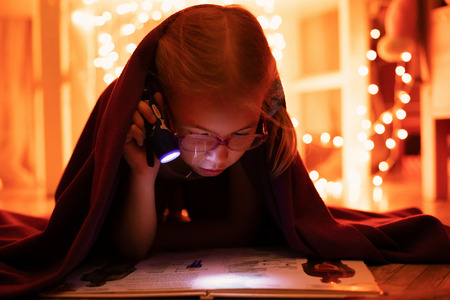 Little blonde girl 7 years old with glasses reading the book under the blanket with little flashlight at dark night time.の写真素材