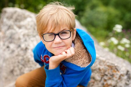 Nice funny blonde boy with strabismus wearing glasses with special lens sits on the stone in the mountains.の写真素材