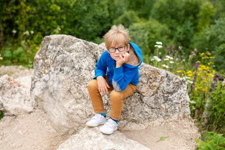 Nice funny blonde boy with strabismus wearing glasses with special lens sits on the stone in the mountains.の写真素材