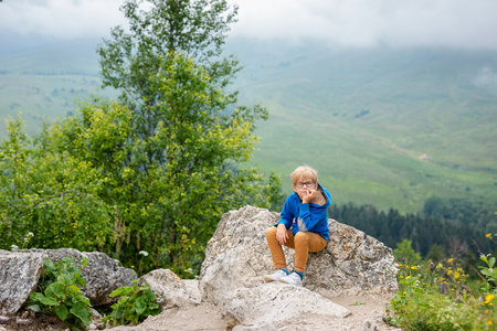 Nice funny blonde boy with strabismus wearing glasses with special lens sits on the stone in the mountains.の写真素材