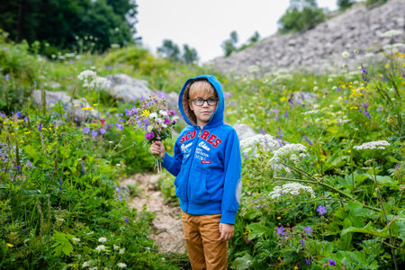 Boy with glasses and blue wear with bouquet of wild flowers in the summer mountains. Flowers for...
