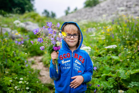 Boy with glasses and blue wear with bouquet of wild flowers in the summer mountains. Flowers for...