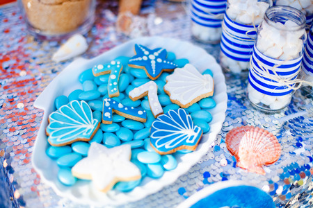 White shell plate full of chocolate candies with blue icing, cookies like shells, stars and number seven and a lot of small jars with marshmallow on the table with shining cloth. Summer season delicious on the party. Sea time theme on the candy bar .の写真素材