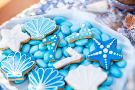 White shell plate full of chocolate candies with blue icing, cookies like shells, stars and number seven and a lot of small jars with marshmallow on the table with shining cloth. Summer season delicious on the party. Sea time theme on the candy bar .の写真素材