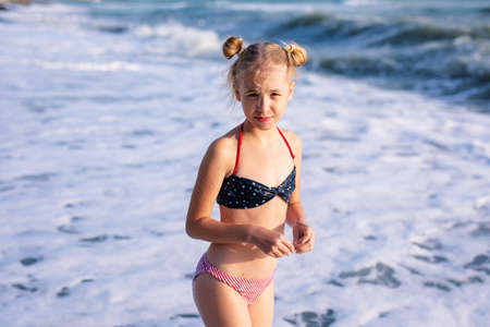 Adorable dissatisfied blonde girl in the white, red and blue swimming suit on beach vacation. Blue sea with white waves on the background.の写真素材