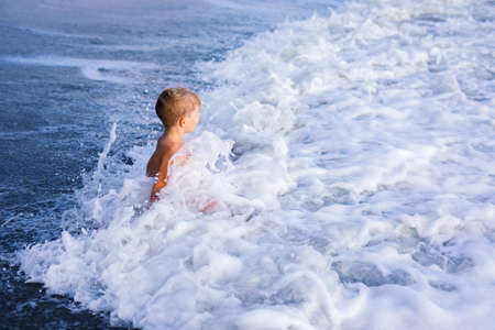 Child boy playing in the waves on the beach in summer sunset, kid watching sea waves and having fun.の写真素材