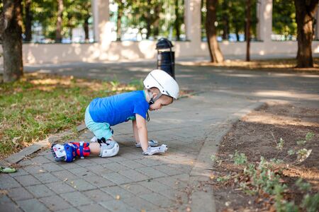 Little blonde boy 3 years old in white sport helmet and blue t-shirt riding on the roller-slates in the summer green park. Special problems with kid's eyes. Myopie, astigmatism, cross-eyed.の写真素材