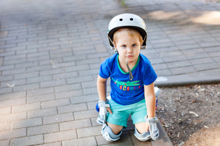 Little blonde boy 3 years old in white sport helmet and blue t-shirt riding on the roller-slates in the summer green park. Special problems with kid's eyes. Myopie, astigmatism, cross-eyed.の写真素材