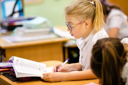 Blonde girl with big glasses sitting in classroom, studing, smiling. Education on elementary school, first day at school.の写真素材
