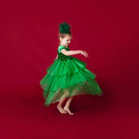 Beautiful little girl princess dancing in luxury green dress isolated on red background. Carnival party with costumes.の写真素材