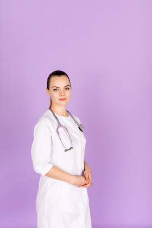 Medical concept of beautiful female doctor in white coat with phonendoscope. Medical student general practitioner. Woman hospital worker looking at camera and smiling, studio, purple backgroundの写真素材