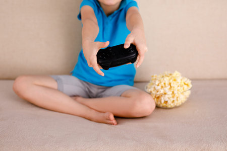 Blonde 9-year-old boy in a blue T-shirt and big glasses sitting on the couch and playing at home...