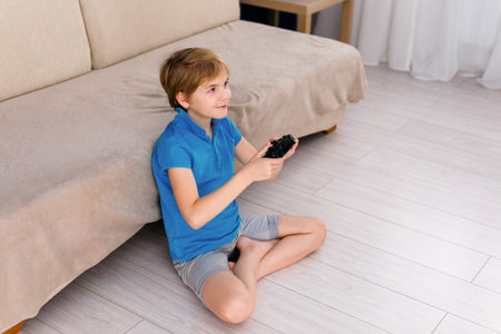 Happy smiling school boy sitting on light couch, playing at home with a gamepad instead of doing his...