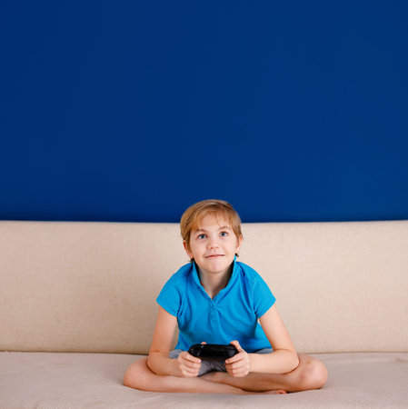 Happy smiling school boy sitting on light couch, playing at home with a gamepad instead of doing his...