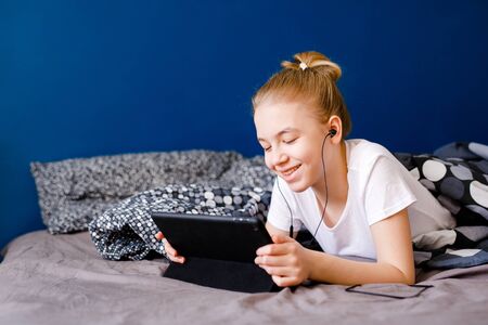 Cute blonde teen girl watching video on smartphone on the bed, child using mobile phone for education. Stay at home quarantine coronavirus COVID-19 pandemic preventionの写真素材