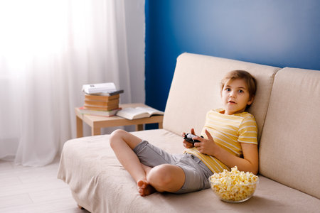Happy smiling school boy in yellow t-shirt sitting on light couch, playing video games with a gamepad instead of doing his homework at home and eating pop corn. Blue background and free space for text.の写真素材