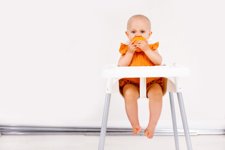 Infant girl sitting in high child's chair eating fruits on a white background. Baby food concept, space for textの写真素材