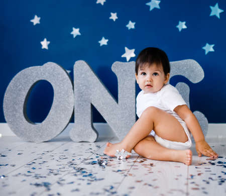One year old baby boy in white t-shirt celebrates birthday near silver letters ONE on blue background with stars and silver sparklesの写真素材