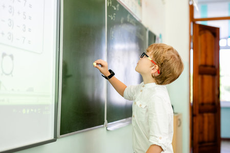 Little boy with big black glasses and white shirt standing near school blackboard with a piece of...