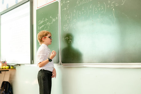 Elementary school student in white shirt and with black glasses writing maths answer on chalkboard